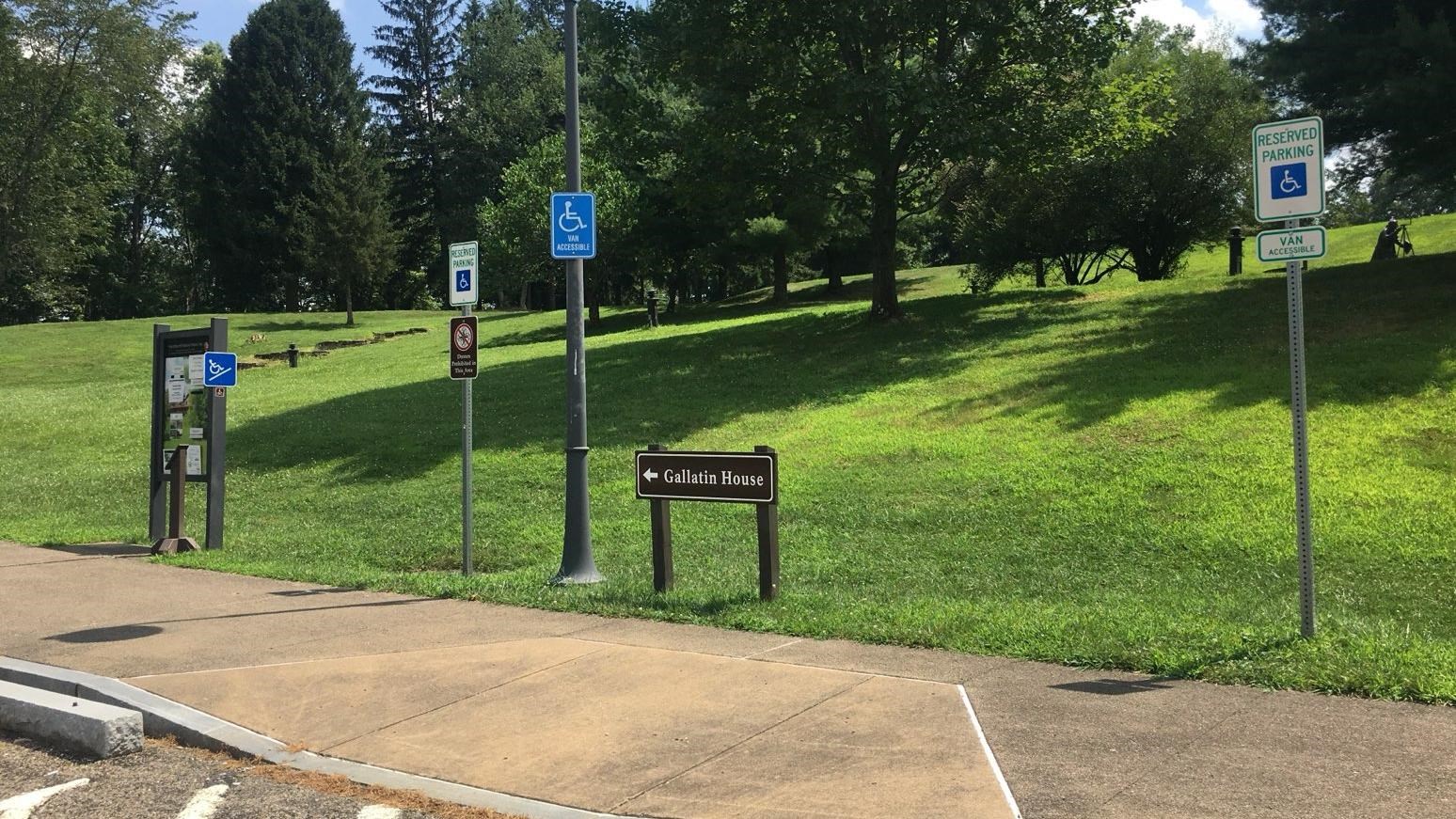 Shows bulletin board and several signs on a green hill. Sidewalk leads up the hill