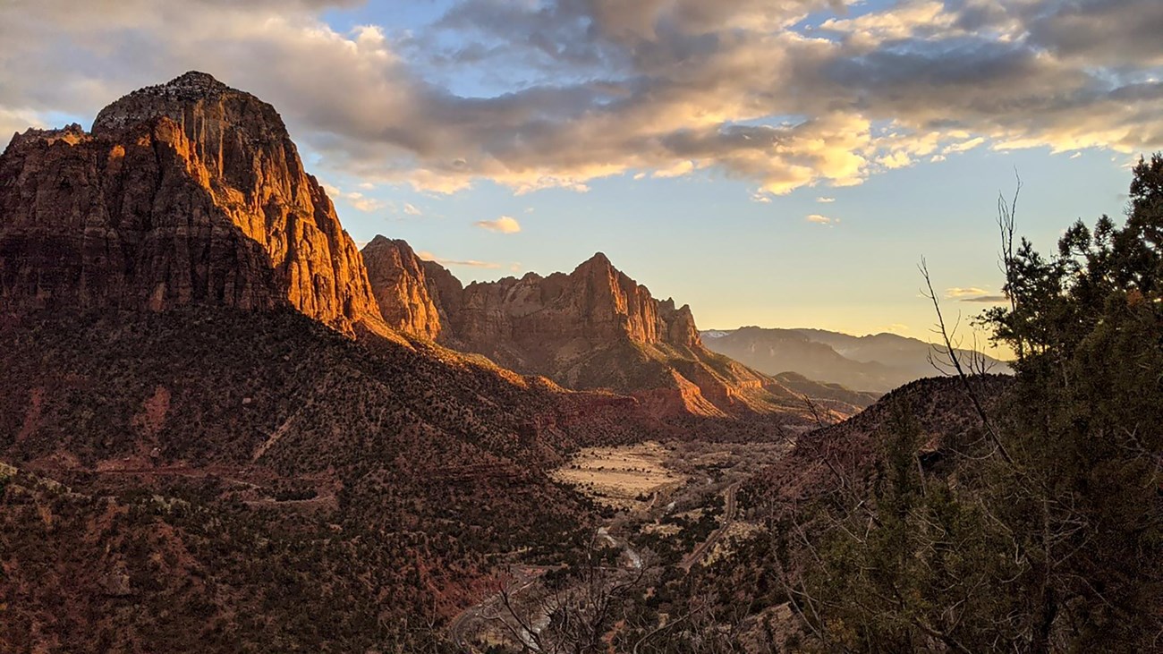 A cloudy blue sky over red sandstone mountains, with green foliage dotting their foothills.