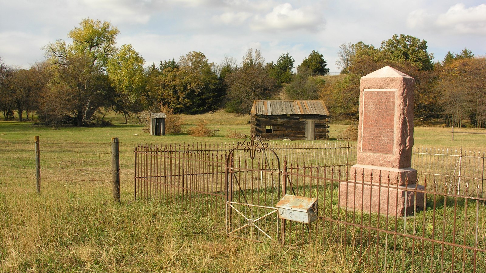 A stone monument sits within a fence in front of a historic log cabin in the distance.