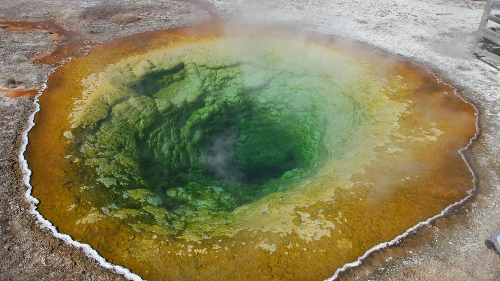 A green, circular hot spring with yellow and orange microbial mats along the edges.