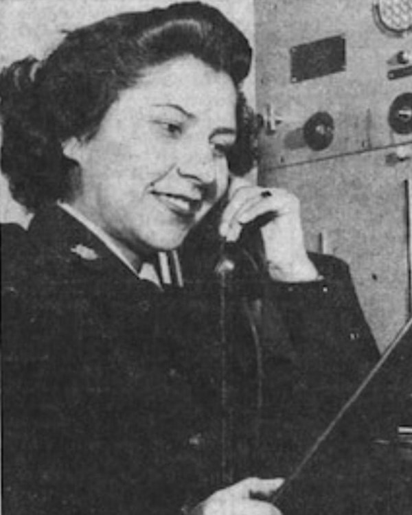 Black and white photo of woman in dark uniform holding a phone with control board in the background