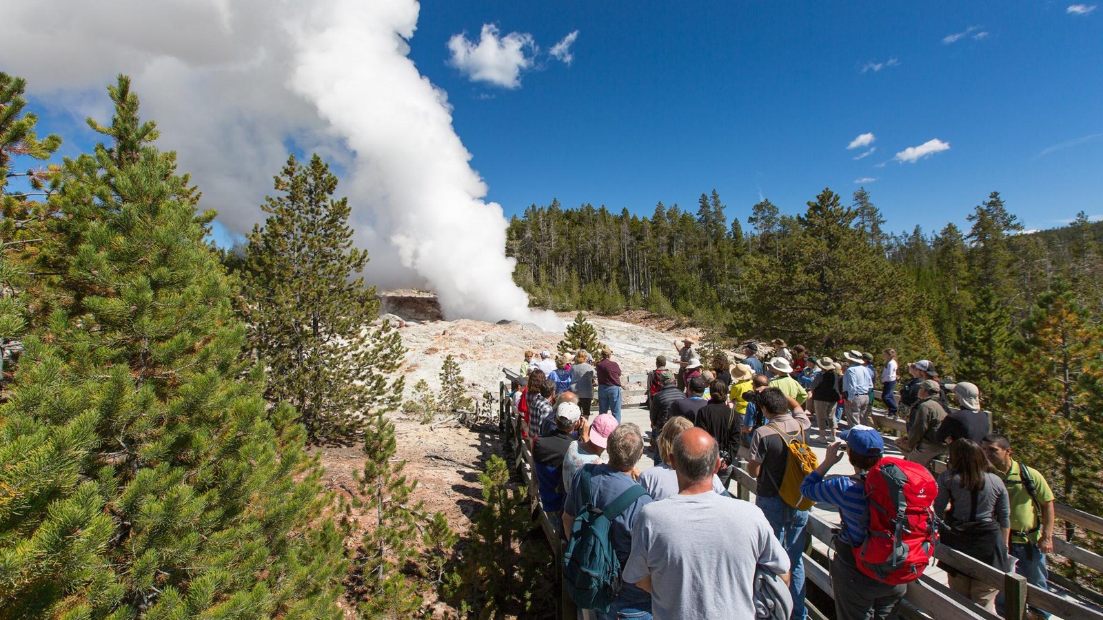 A crowd of people gather on a viewing platform and watch a large steam plume from a geyser.