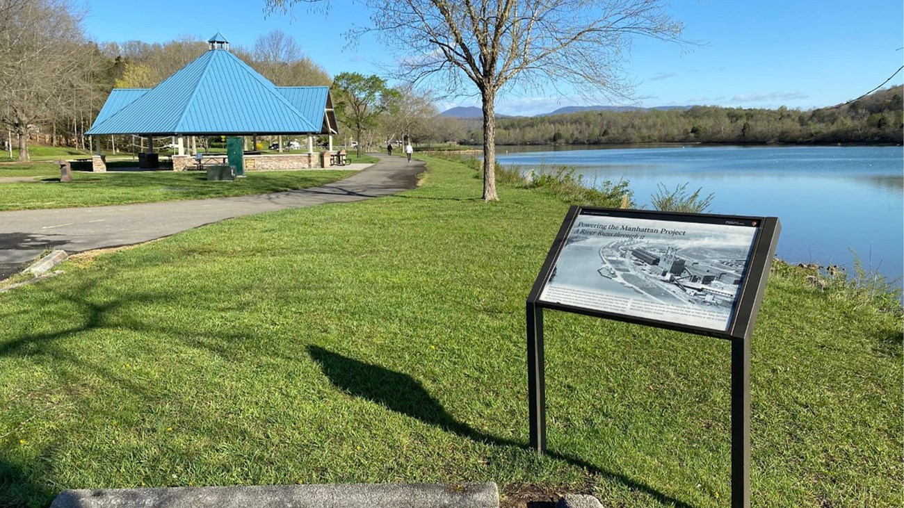 An historical marker on the bank of a calm river with a paved trail and pavilion in the background. 