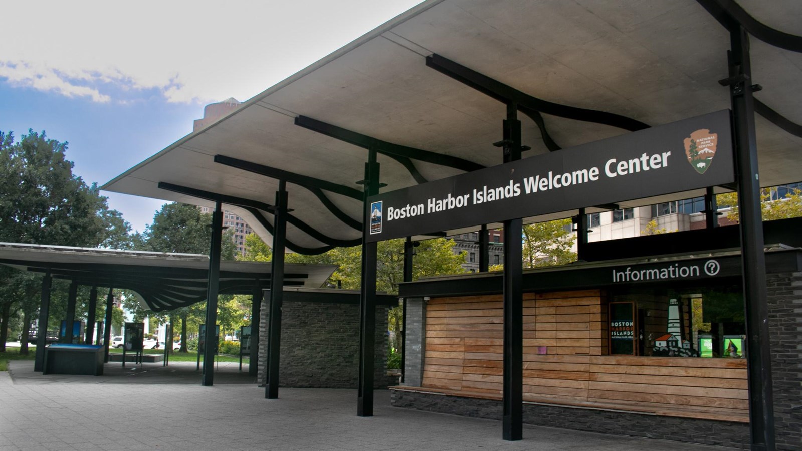 A pavilion hangs over green space, pavement, and the visitors center. The center has wood panelling.