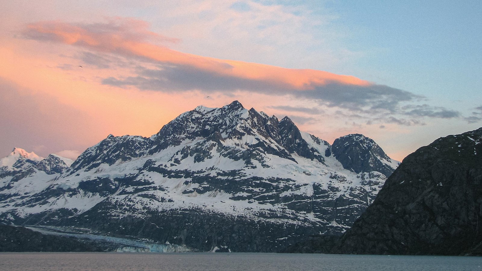 Rugged mount cooper, partially snowcovered, rises high above water and a glacier beneath it.