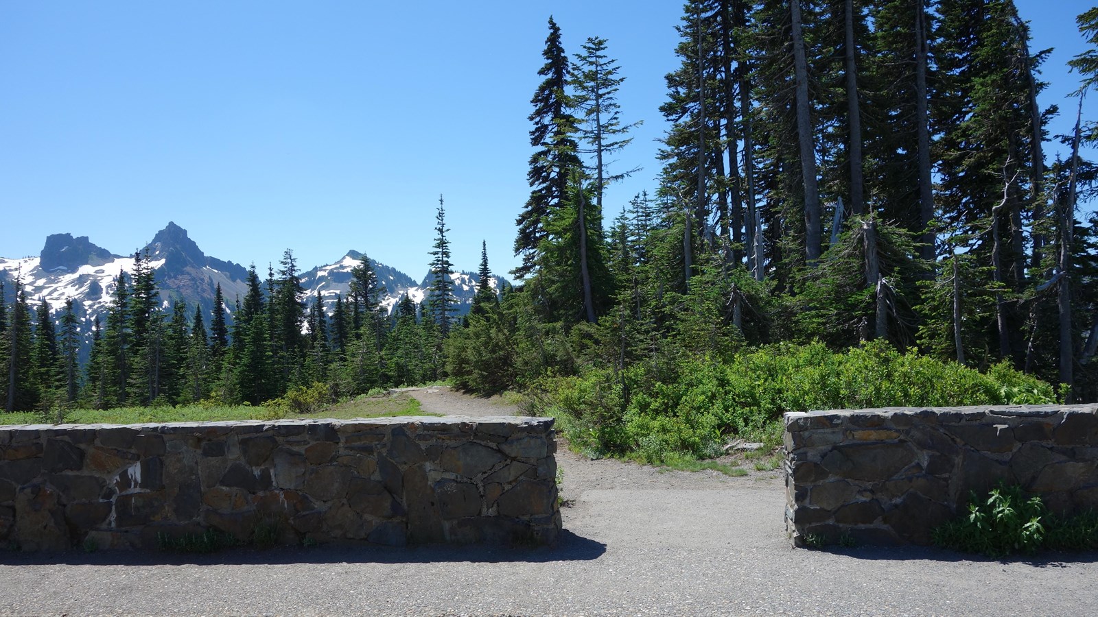 A trail starts from a break in a low rock wall bordering a parking area.