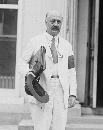 black and white photo of a bald man with a mustache stands in a light colored suit. 