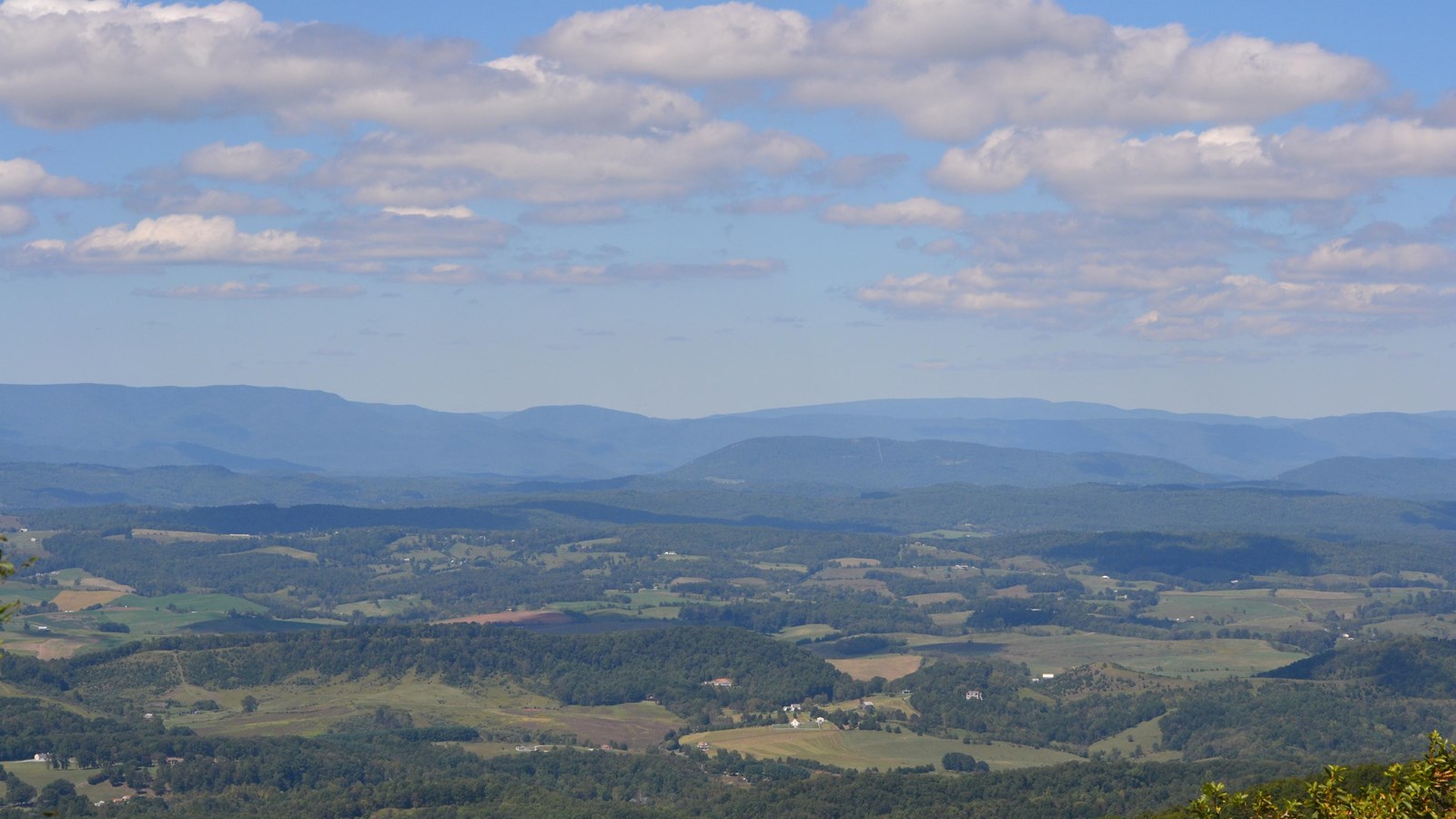 Below, a valley with a few houses and distant, round-topped mountains on the horizon. Fluffy clouds.