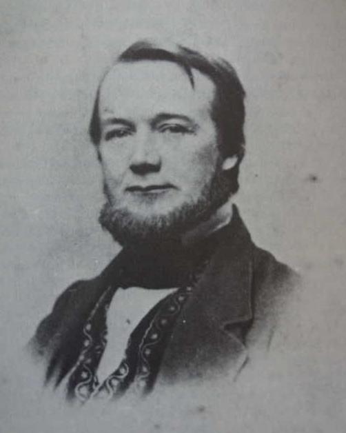 Portrait of a man wearing a dark suit with printed vest underneath. He has a short beard. 