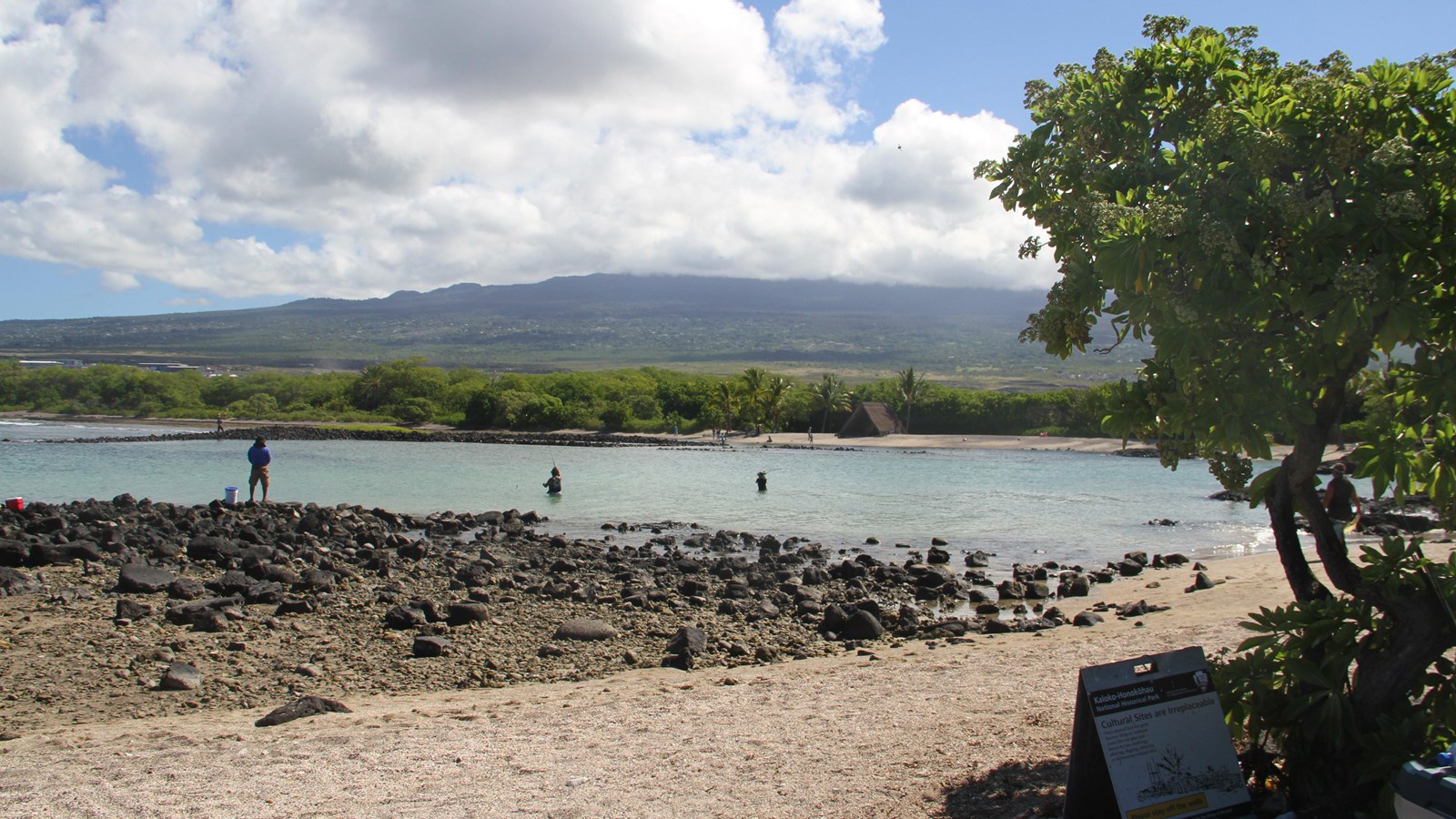 White sand and black lava in foreground, backed by a shallow bay and a distant cloud-covered volcano