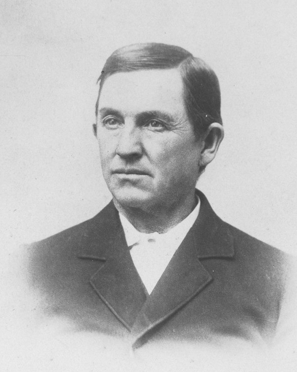 A late 1800s portrait photo depicts a man with a coat and white collared shirt. 