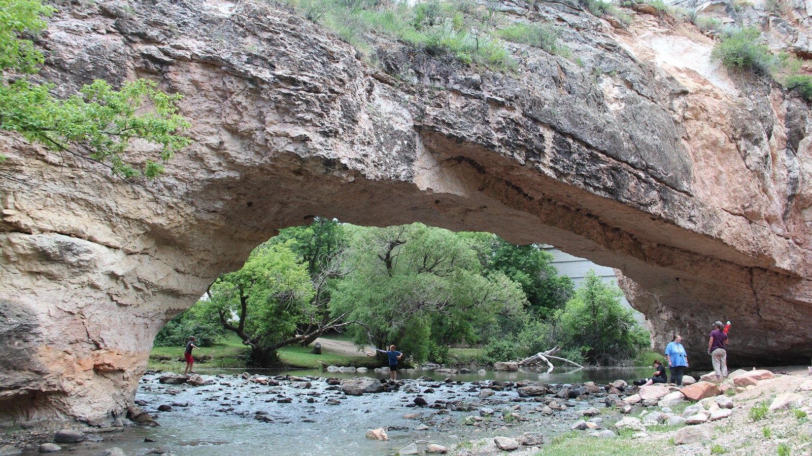 Shallow creek flows under red stone bridge with people exploring the scenic spot.