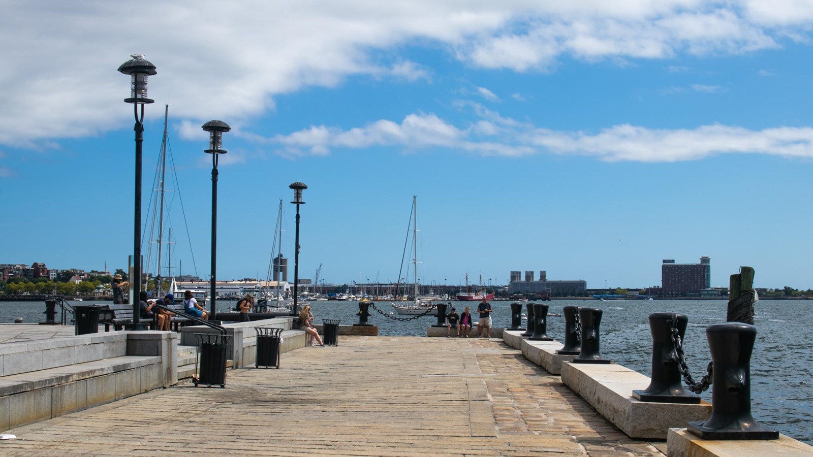 A long stone walkway leads to the edge of the water in the harbor, with a sailboat in the harbor. 