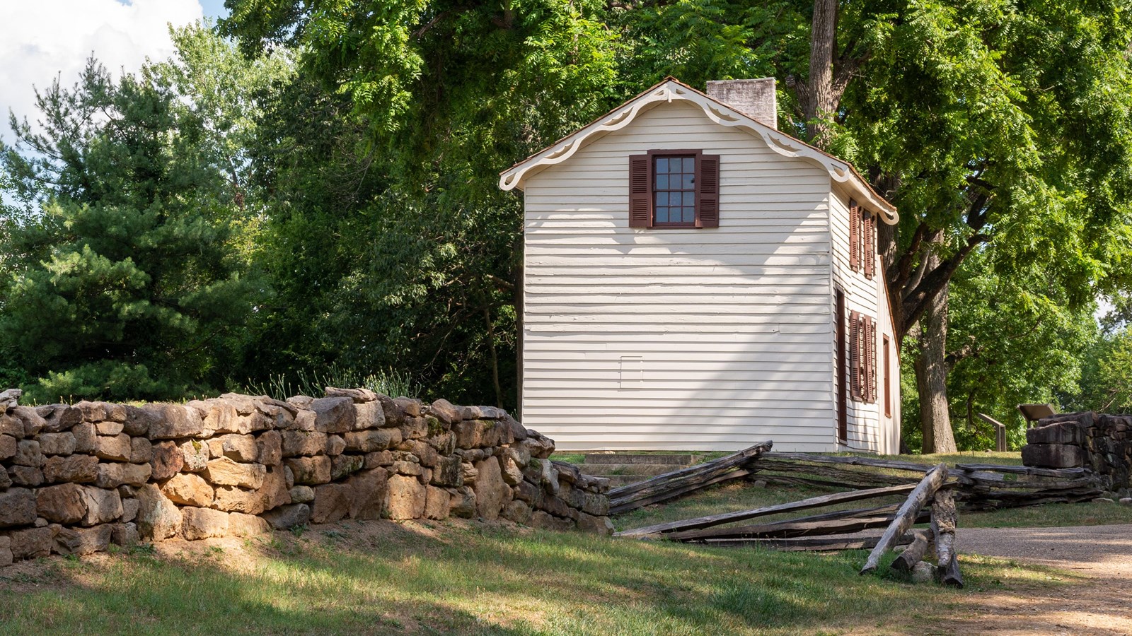 A small, two story white house along a gravel road with a stone wall along it.