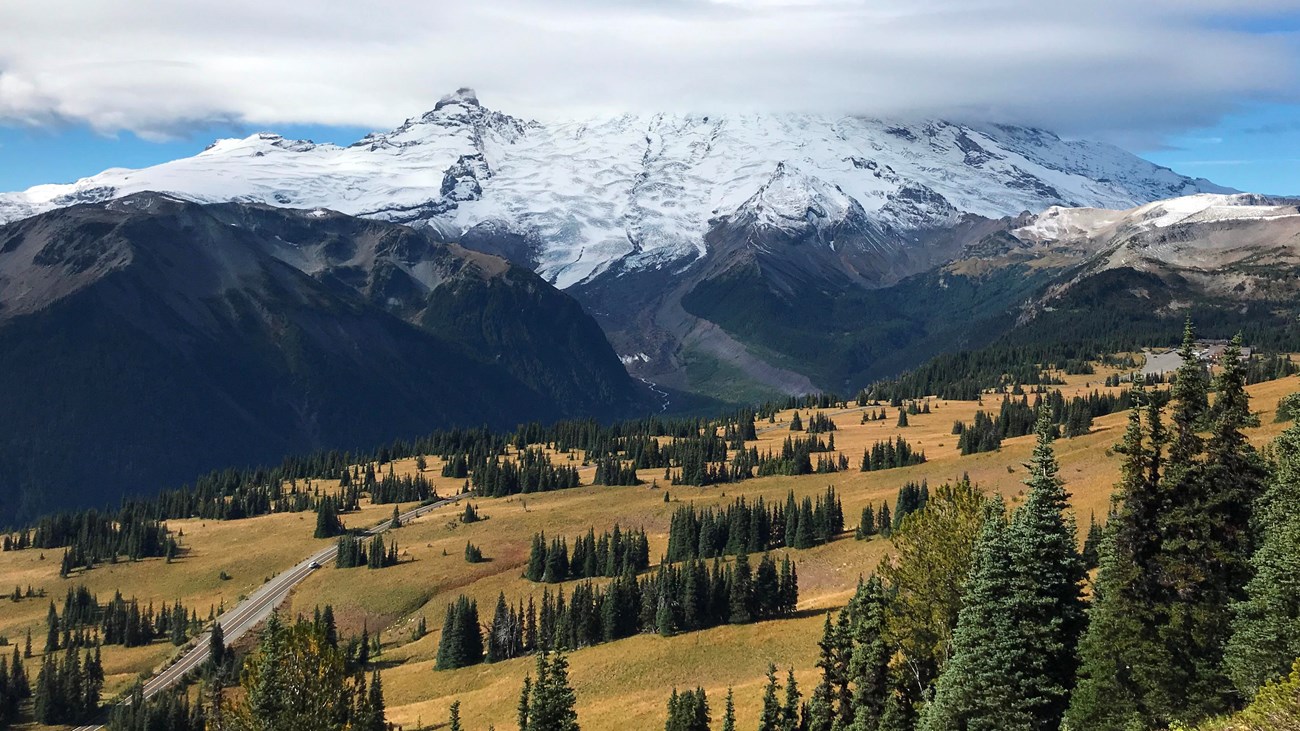 A road curves through subalpine meadows underneath a glaciated peak wrapped in clouds. 