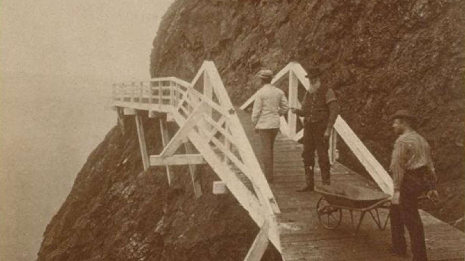  Man holding a wheel barrow, next to two others on a wooden walkway attached to a cliff.