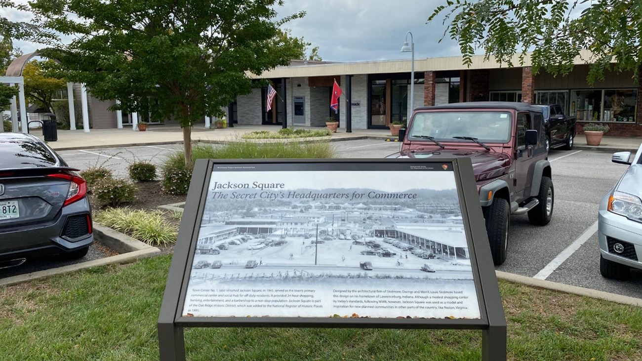 A strip mall with cars parked and historical marker in the foreground.