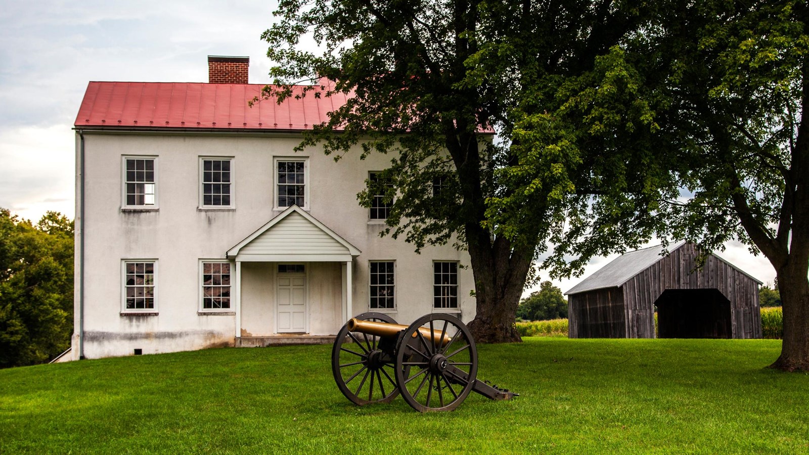 A two-story white farmhouse with a cannon in front and a farm building to the right.