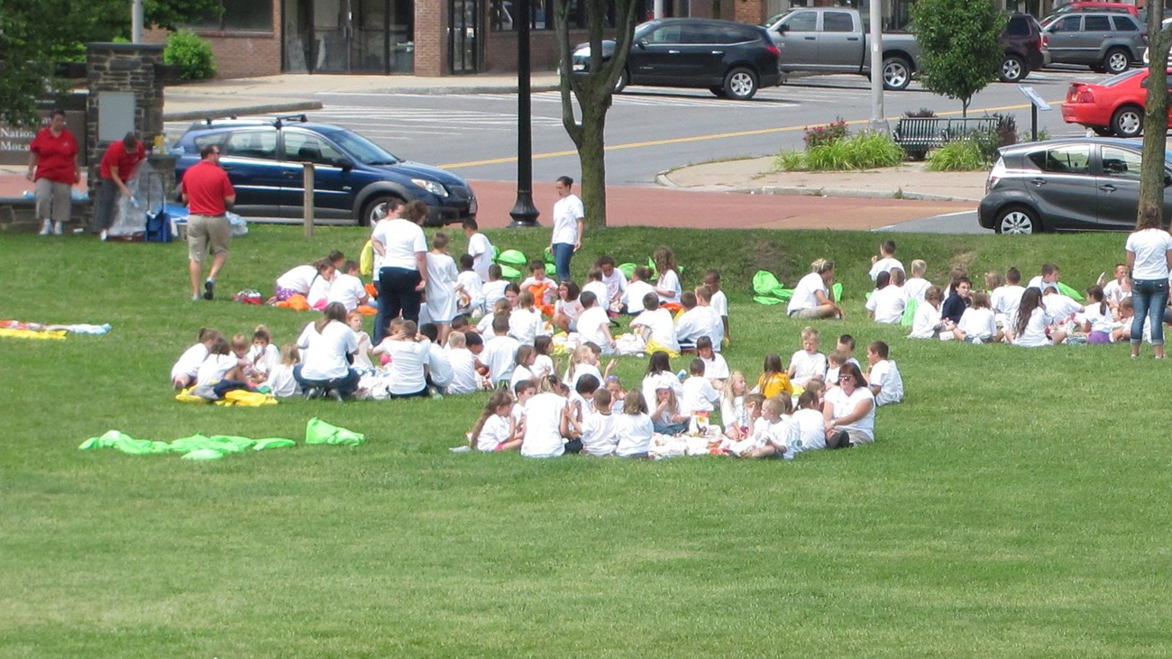 A large group of children sit on blankets scattered across the lawn.
