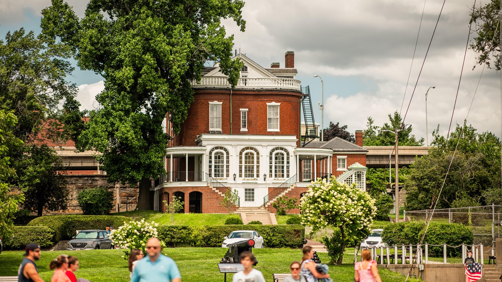 A large brick building with white wooden accents sits amongst a large yard as visitors walk in front