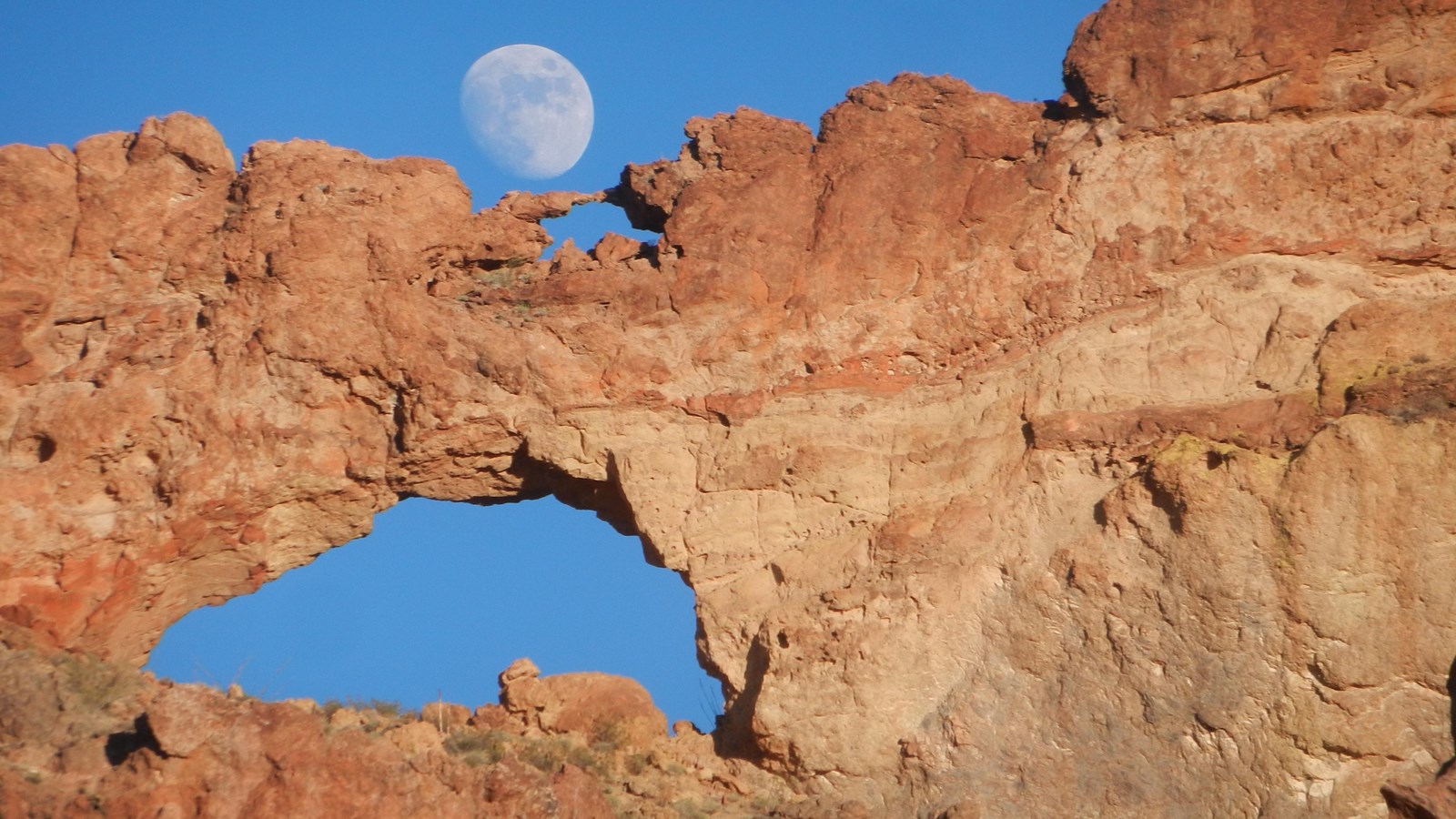 The moon rises behind two natural rock arches, stacked upon one another. 