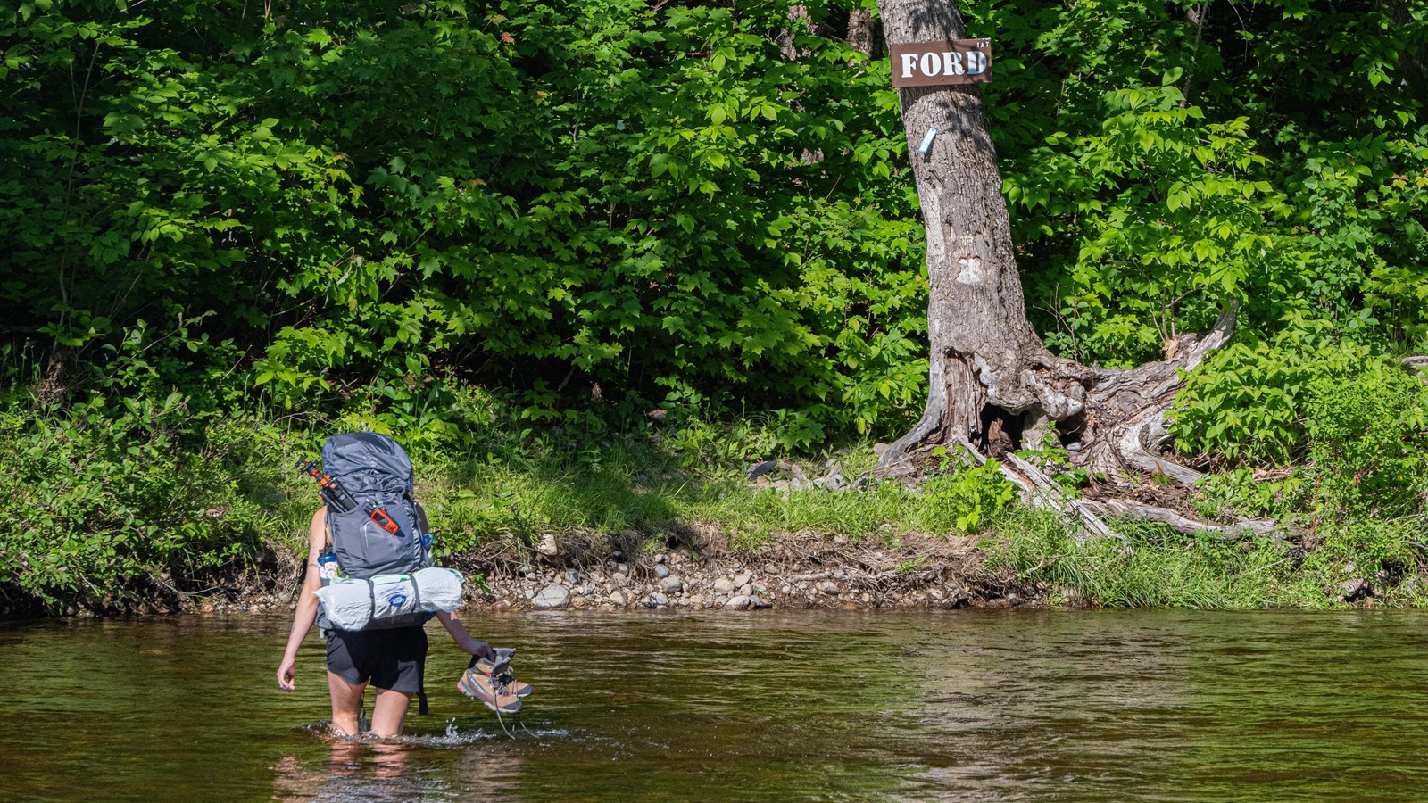 A backpacker in thigh deep water crosses a stream to the opposite shore.