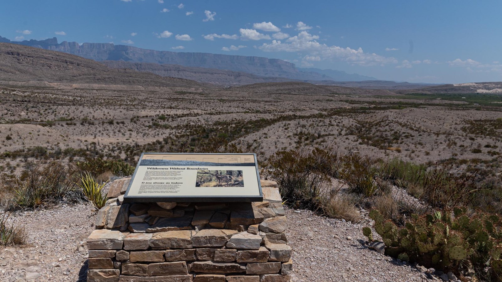 A metal sign sits on a stone pedestal at on overlook that provides a great view of distant mountains