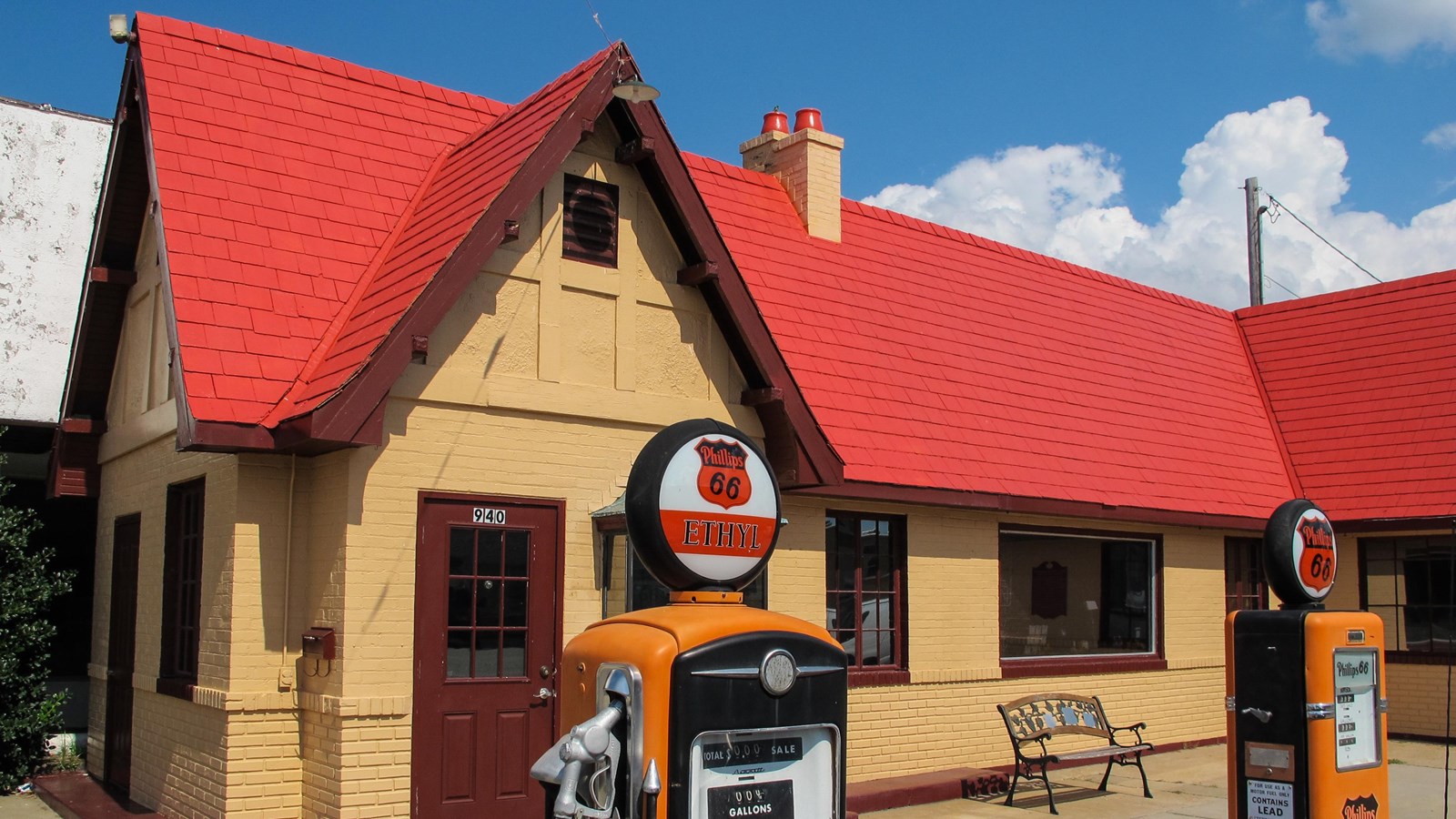 A tan building with a steep red gabled roof. Out front are two yellow and black gas pumps.