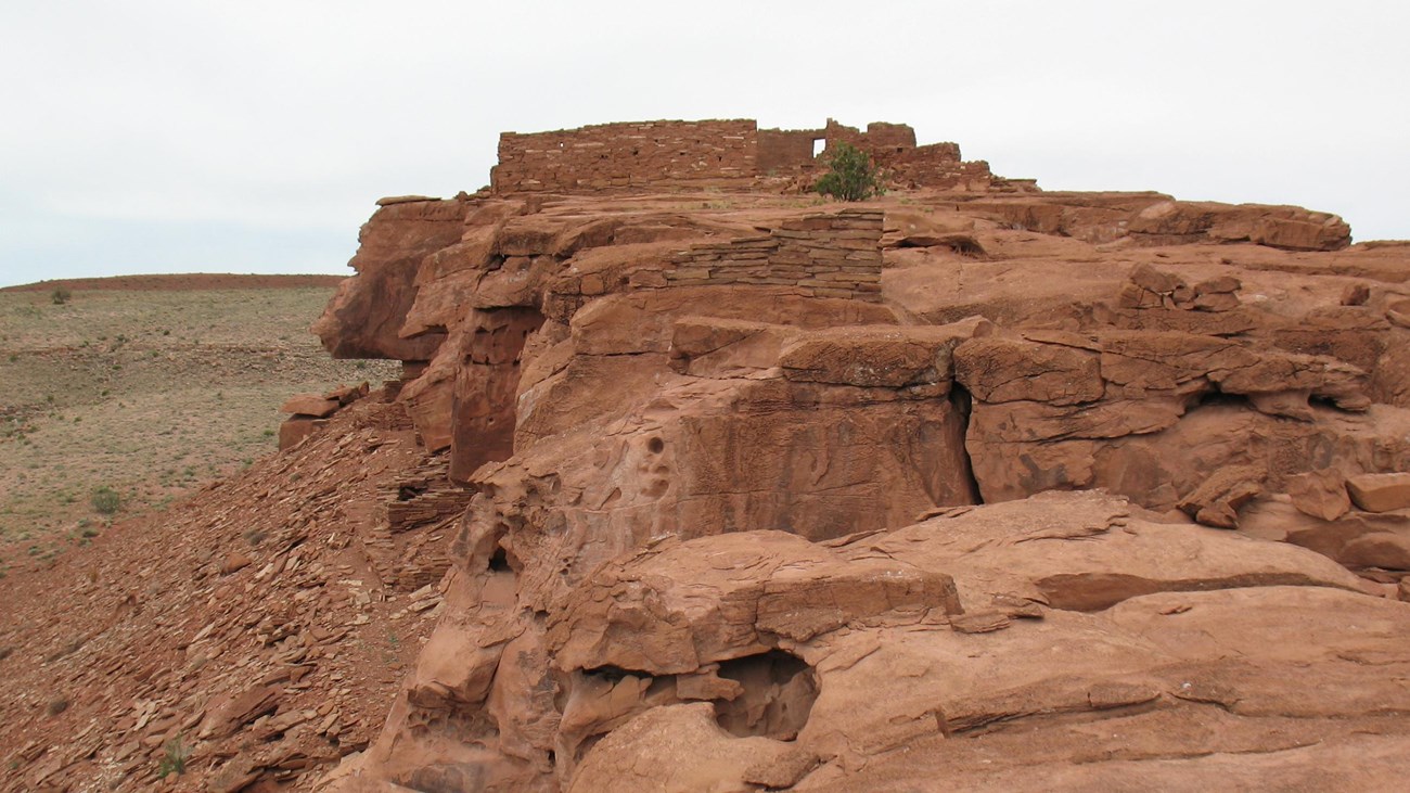 A partial sandstone pueblo on top of a rock outcrop with grassy hills in the distance. 