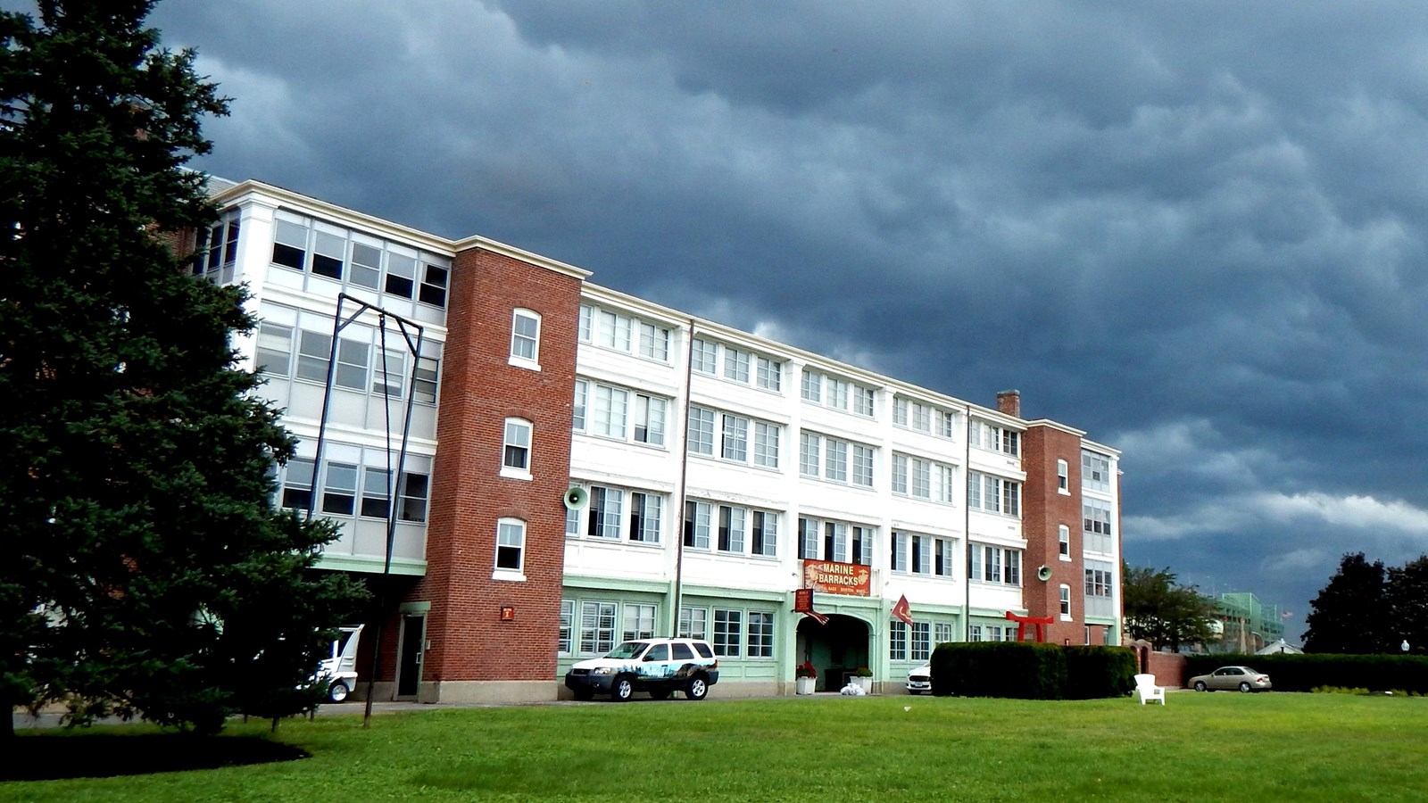 Cloud-filled sky over the Marine Barracks, a four-story building with a grassy field in front.