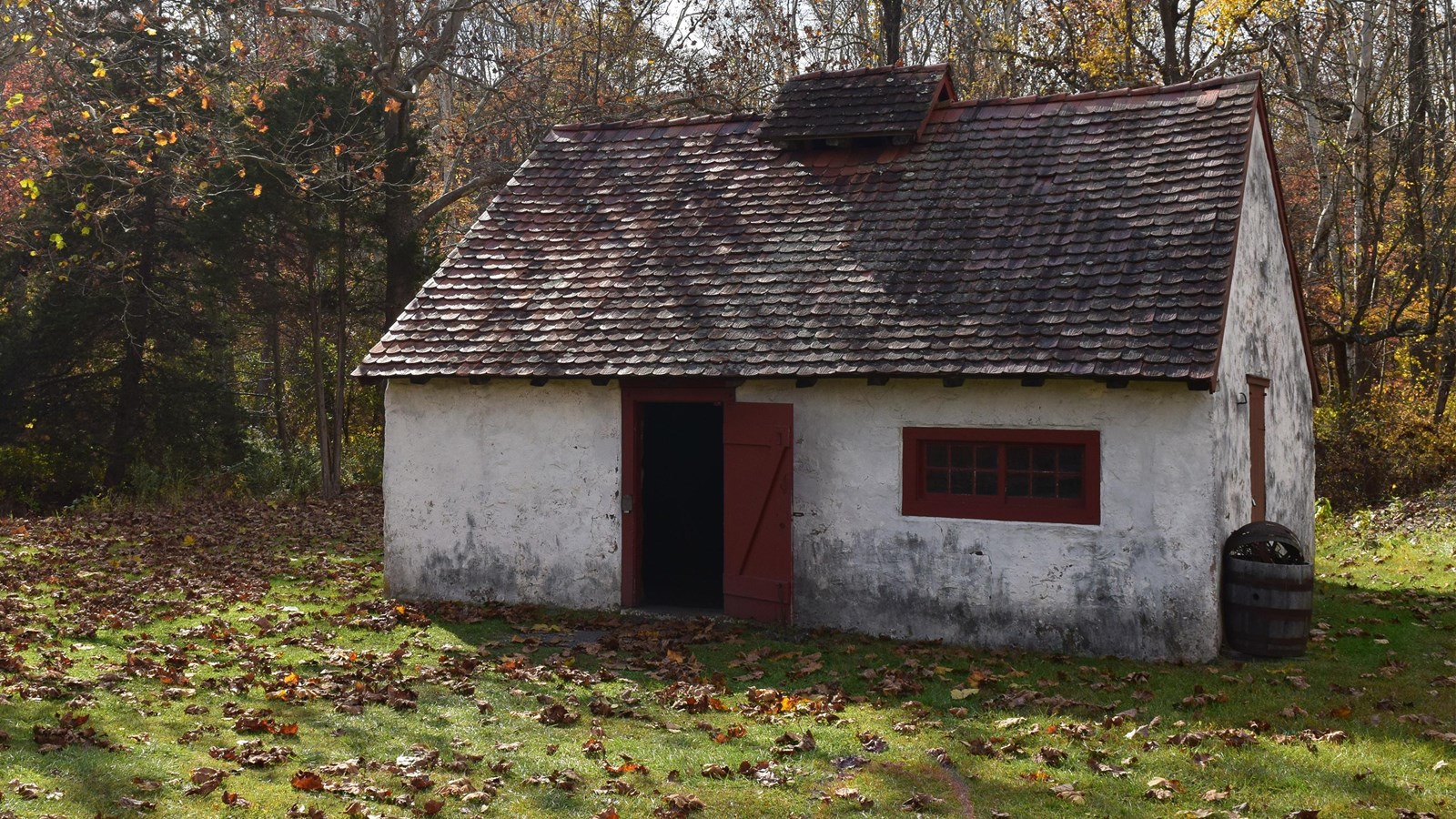 a white stone building with a red roof and an open door. A rusted barrel sits next to the building. 