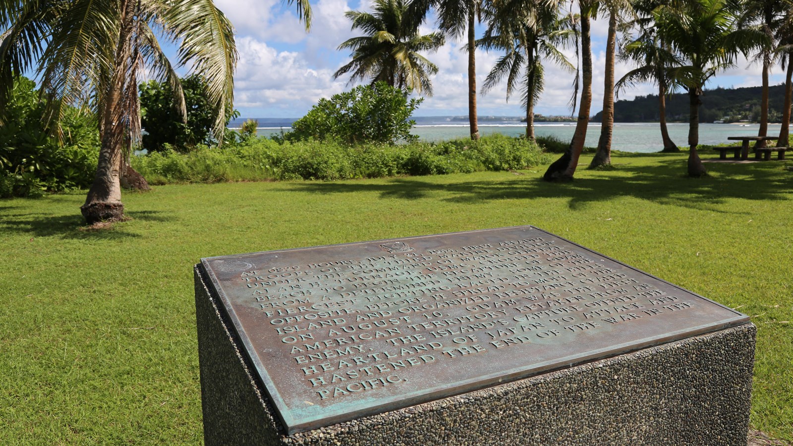 Bronze plaque set on a concrete cube. In the background are palm trees and the ocean.