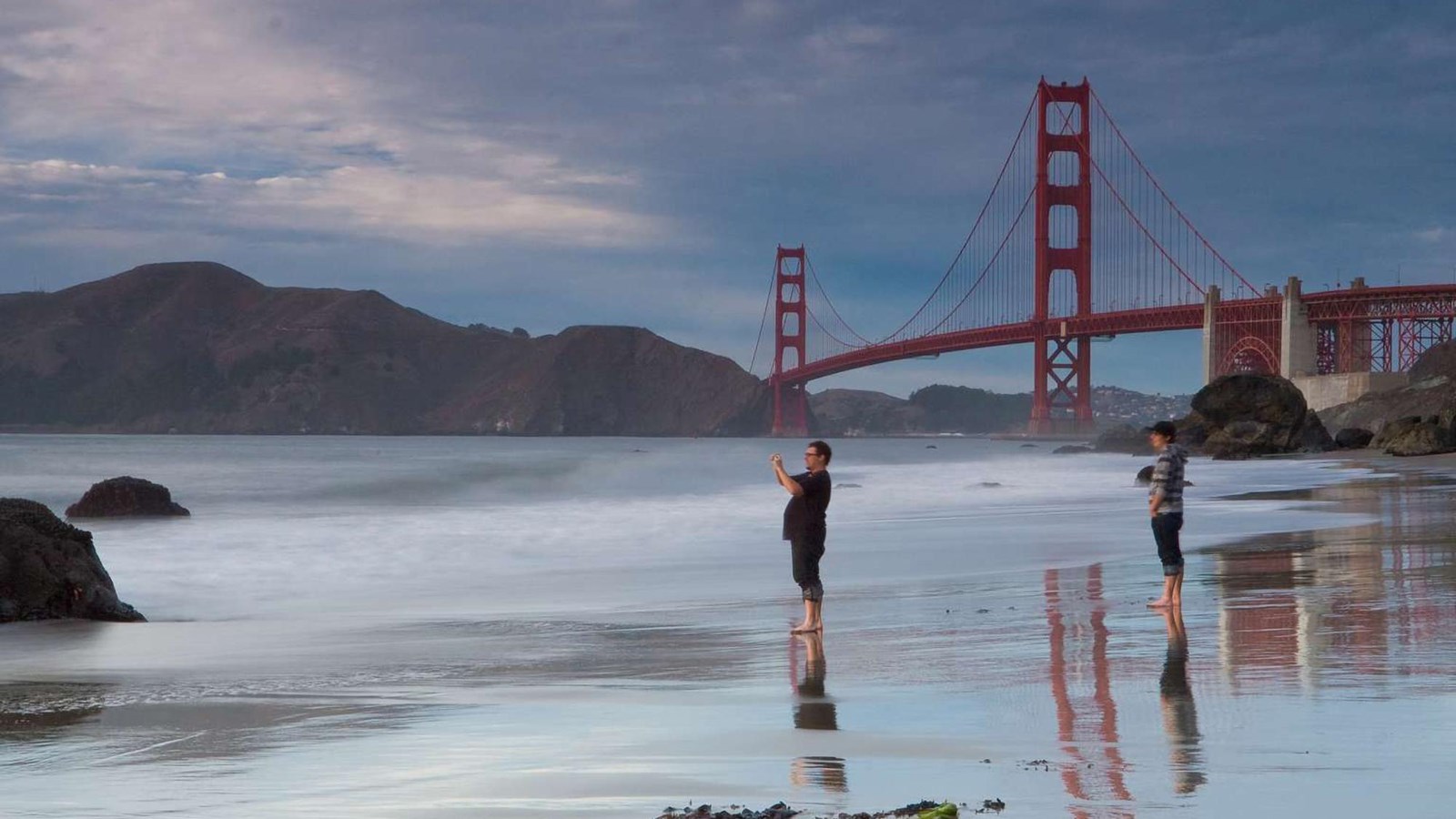 Visitors stand barefoot on the sand at dusk, the Golden Gate Bridge looms in the distance.