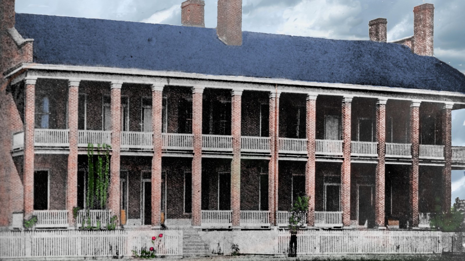 Colorized historic photo 2-story red brick building with porch and balcony running its length. 