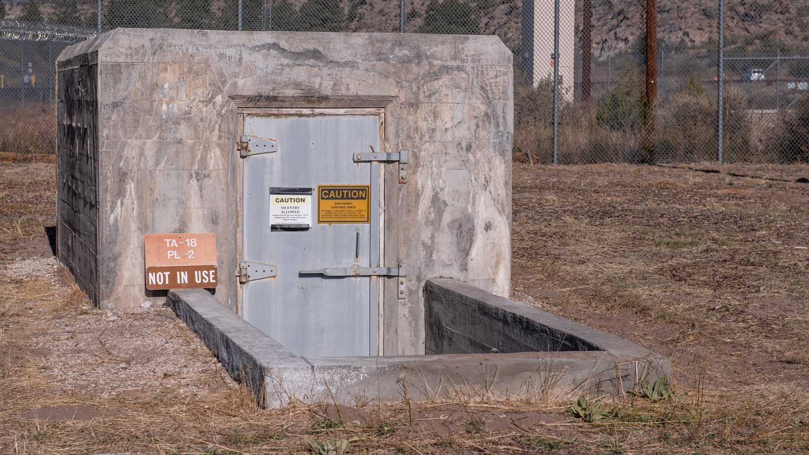 A door set in a concrete frame sits recessed in the ground, with a set of stairs leading down to it.