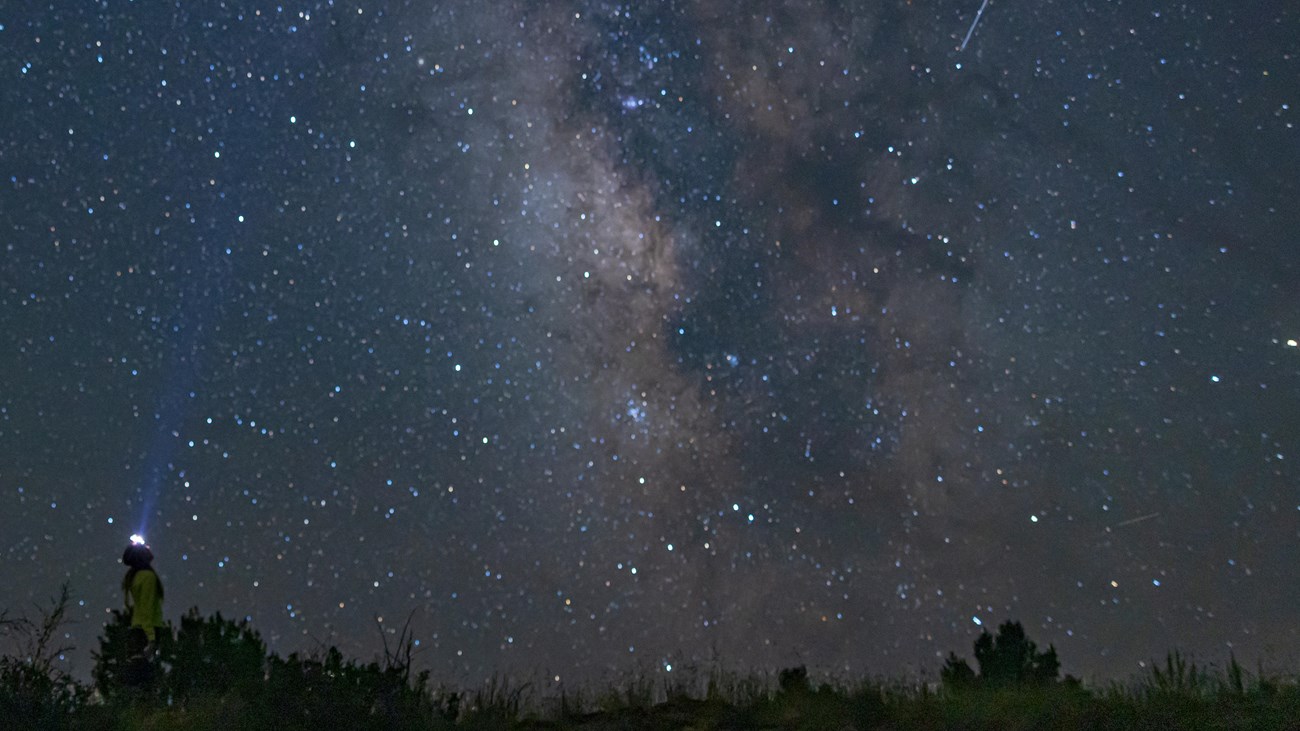 A hiker with a headlamp looks up at the star-filled night sky. The Milky Way Galaxy creates a band o