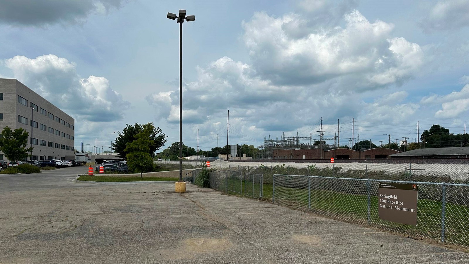 View of a parking lot with a fence on the right side. A sign for the park is posted on the fence.