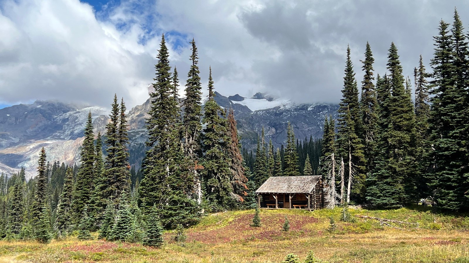 Log cabin surrounded by trees and meadow with clouds and glaciated mountain behind it