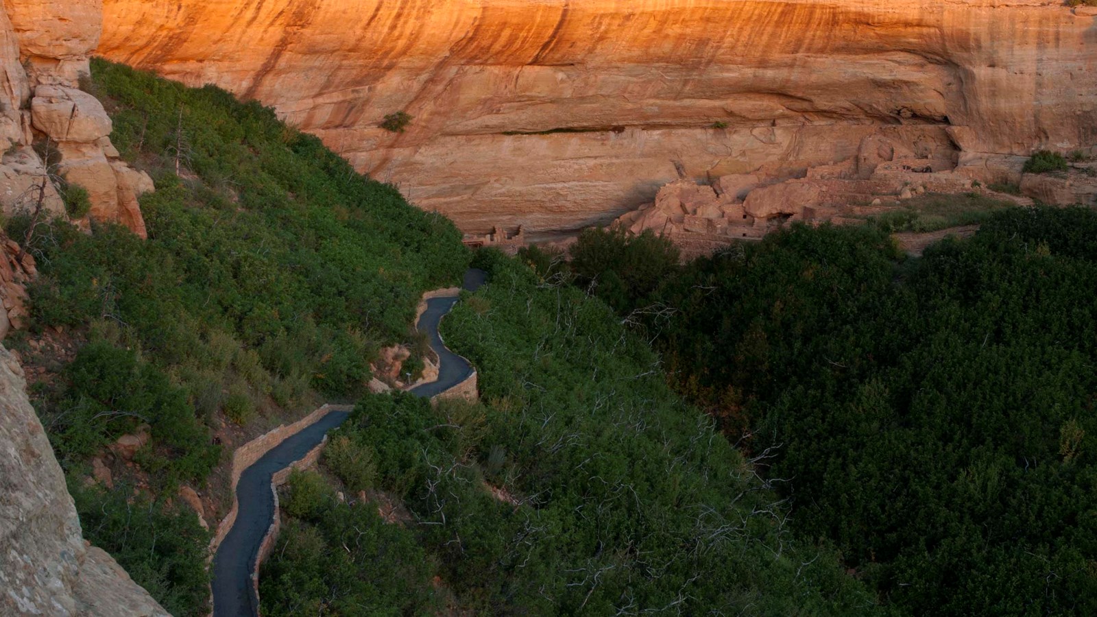 A paved trail winds through scrub oak to a stone-masonry cliff dwelling built in a sandstone alcove 