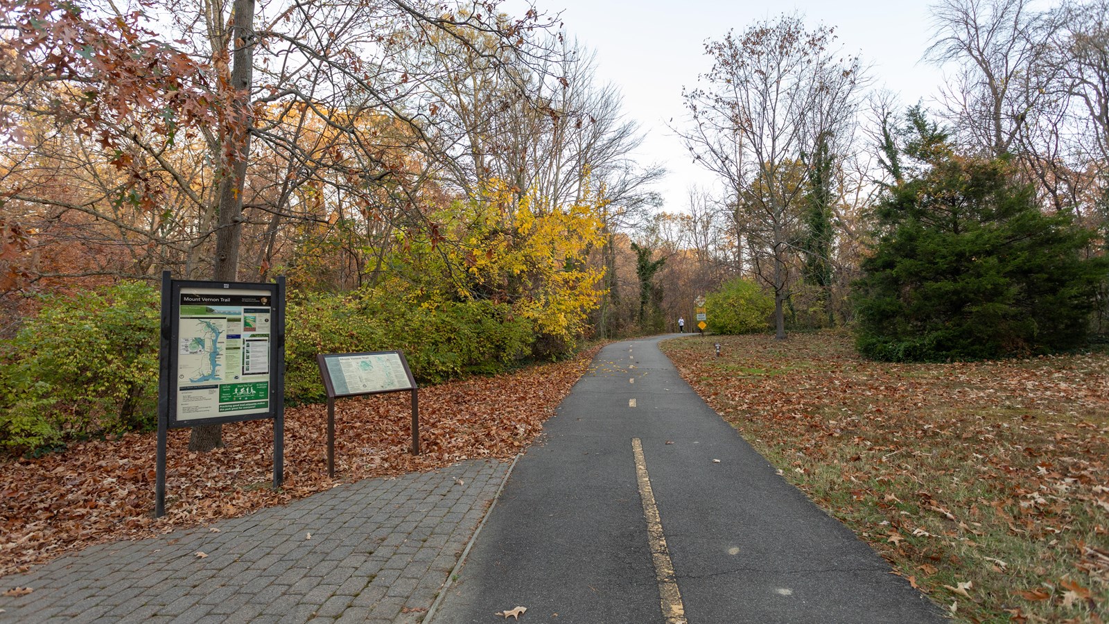 A paved pedestrian walkway is positioned to the right of two information panels. Trees surround.