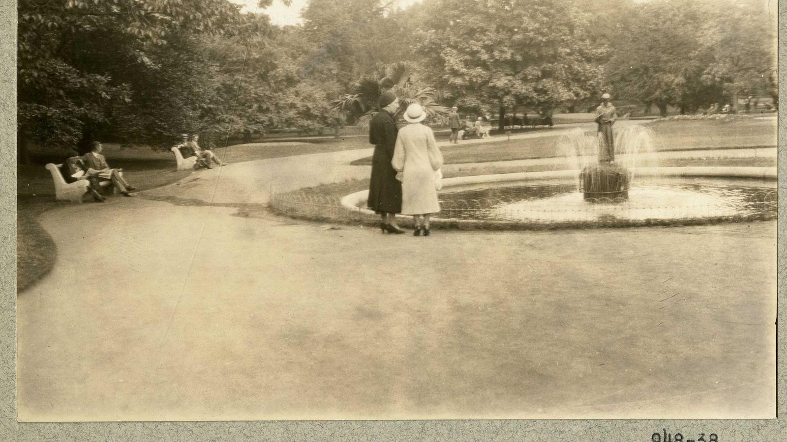 Black and white of path going around fountain with people on it, benches and trees on side