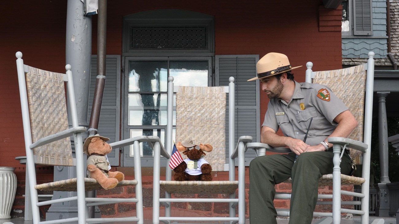 A Park Ranger sits in a rocking chair next to a stuffed moose toy and a stuffed teddy bear. 