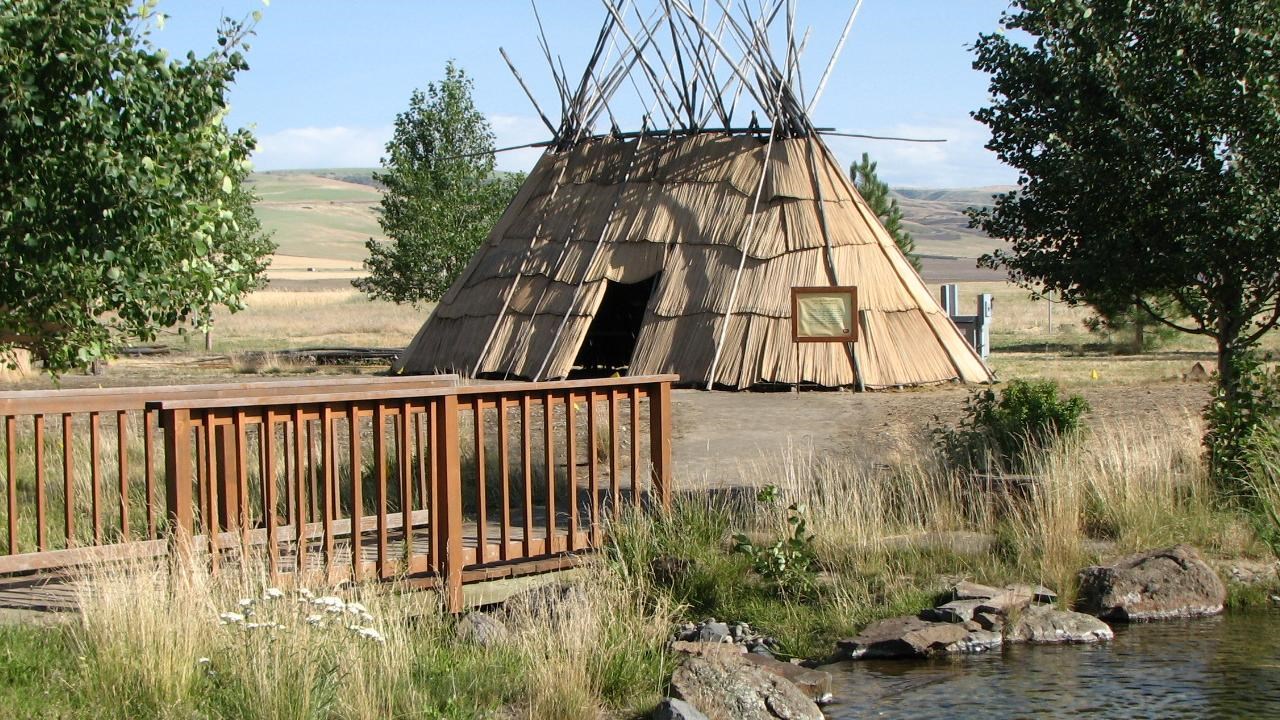 Large hut structure made of tule reeds and wooden posts with rolling prairie hills