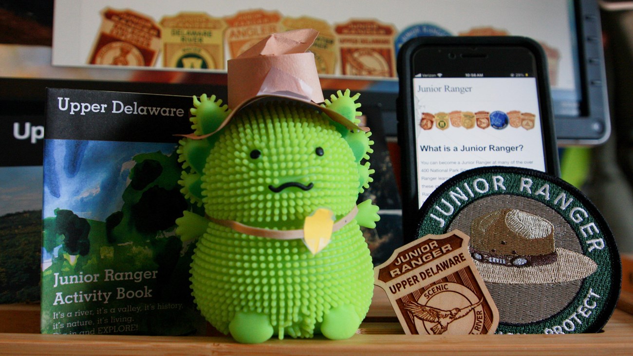A toy axolotl with a paper ranger hat/badge next to Upper Delaware Junior ranger badge, patch, book 