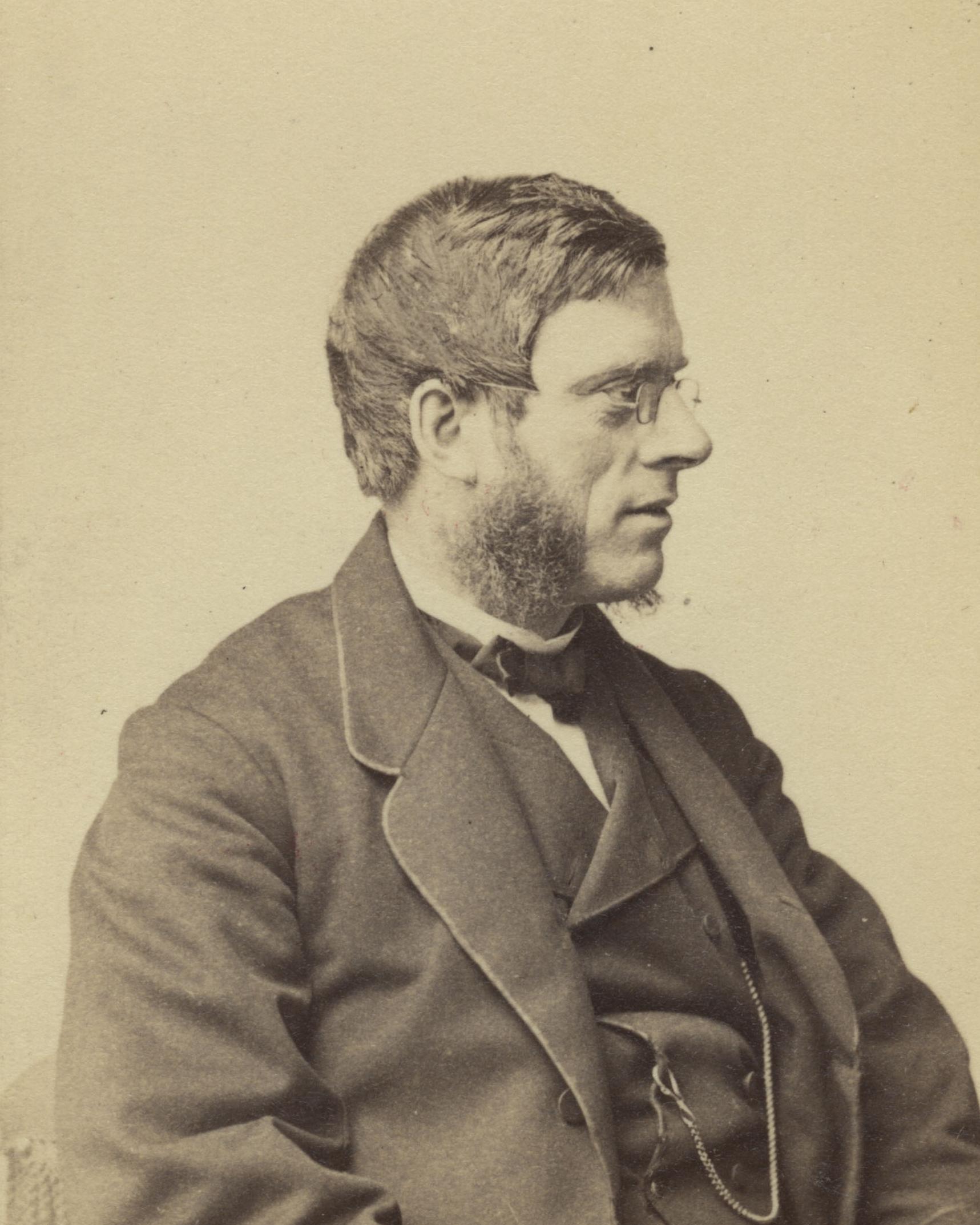 Side sitting portrait of man in a three-piece suit wearing glasses and sideburns
