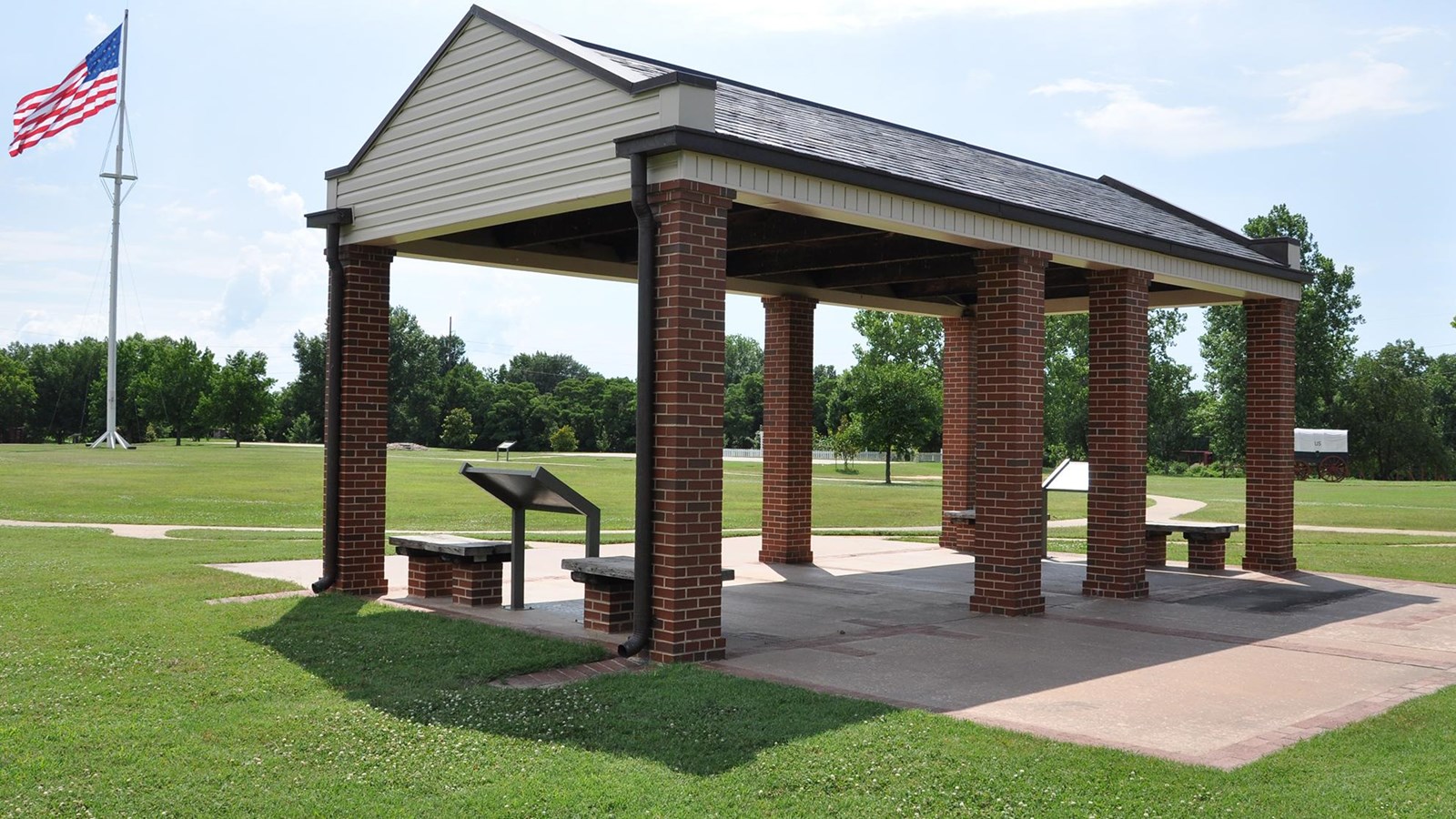 8 column brick pergola covers part of a cement slab surrounded tall flag pole in the background. 