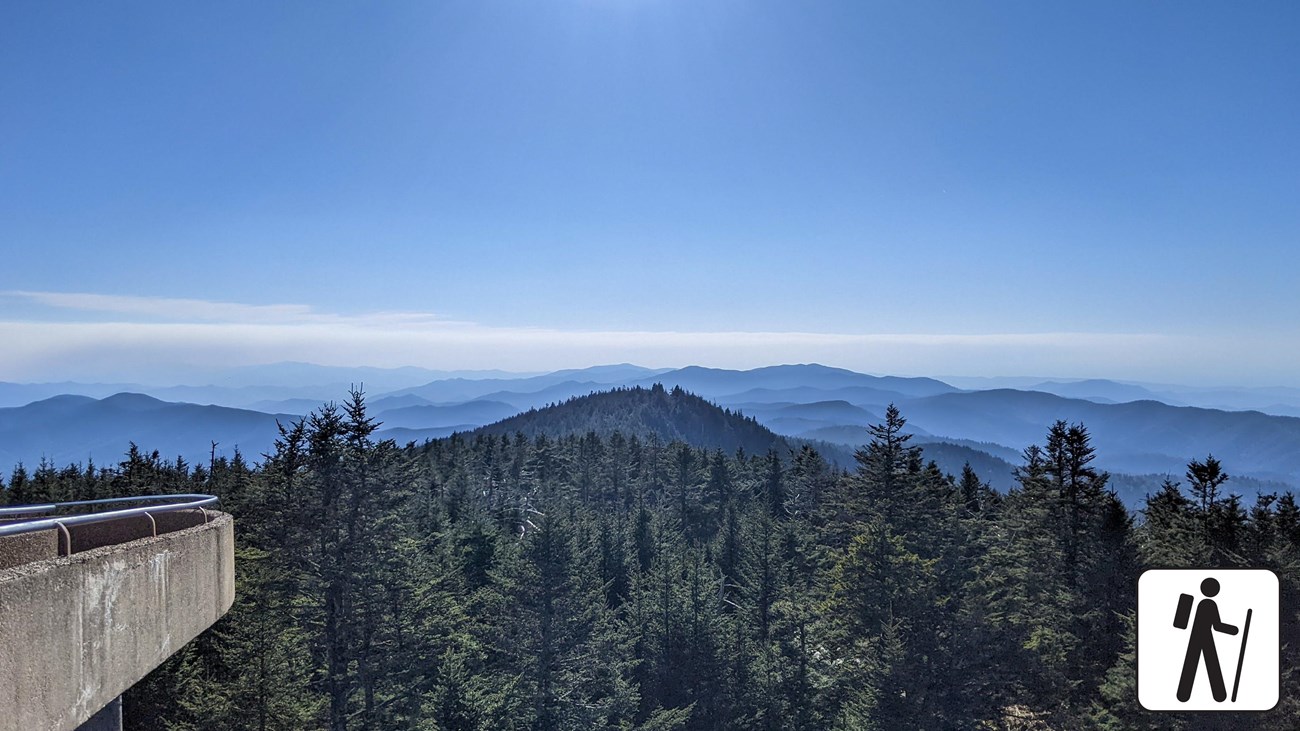 An elevated, paved walkway to a round observation tower overlooking mountains. Hiker icon in corner.