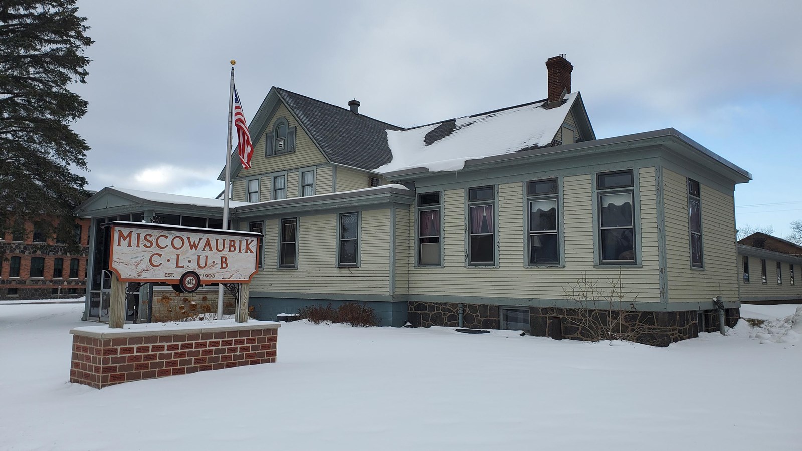 Winter scene of two-story house building with yellow wooden siding and a brick-based sign out front.