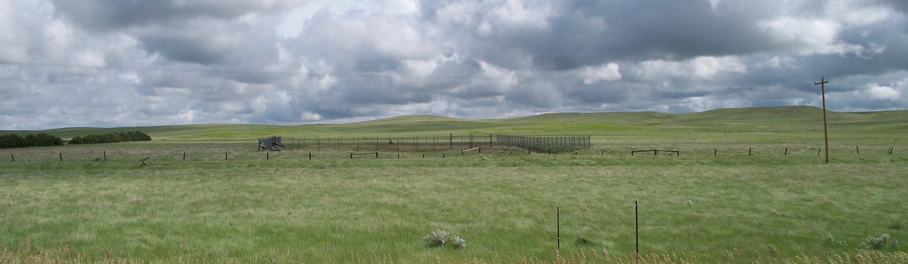 View from a highway into the prairie; in the foreground a rectangular fence breaks up a field.