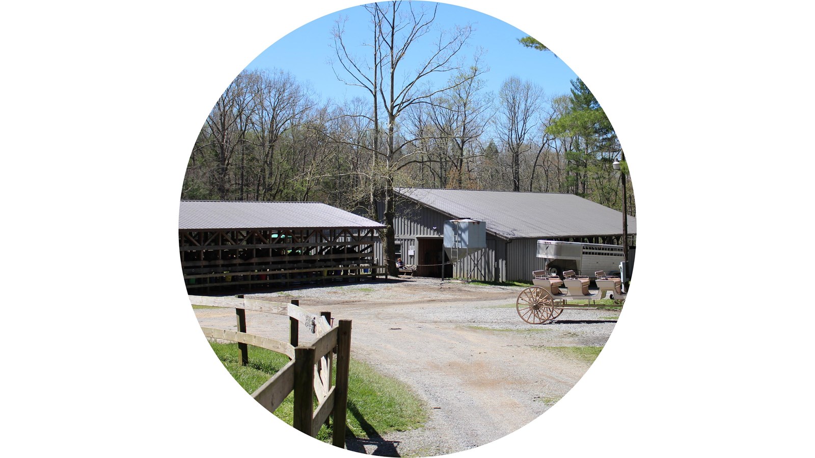 A gravel road leads to a riding stable, barn area, and horse trailer. A carriage in foreground.